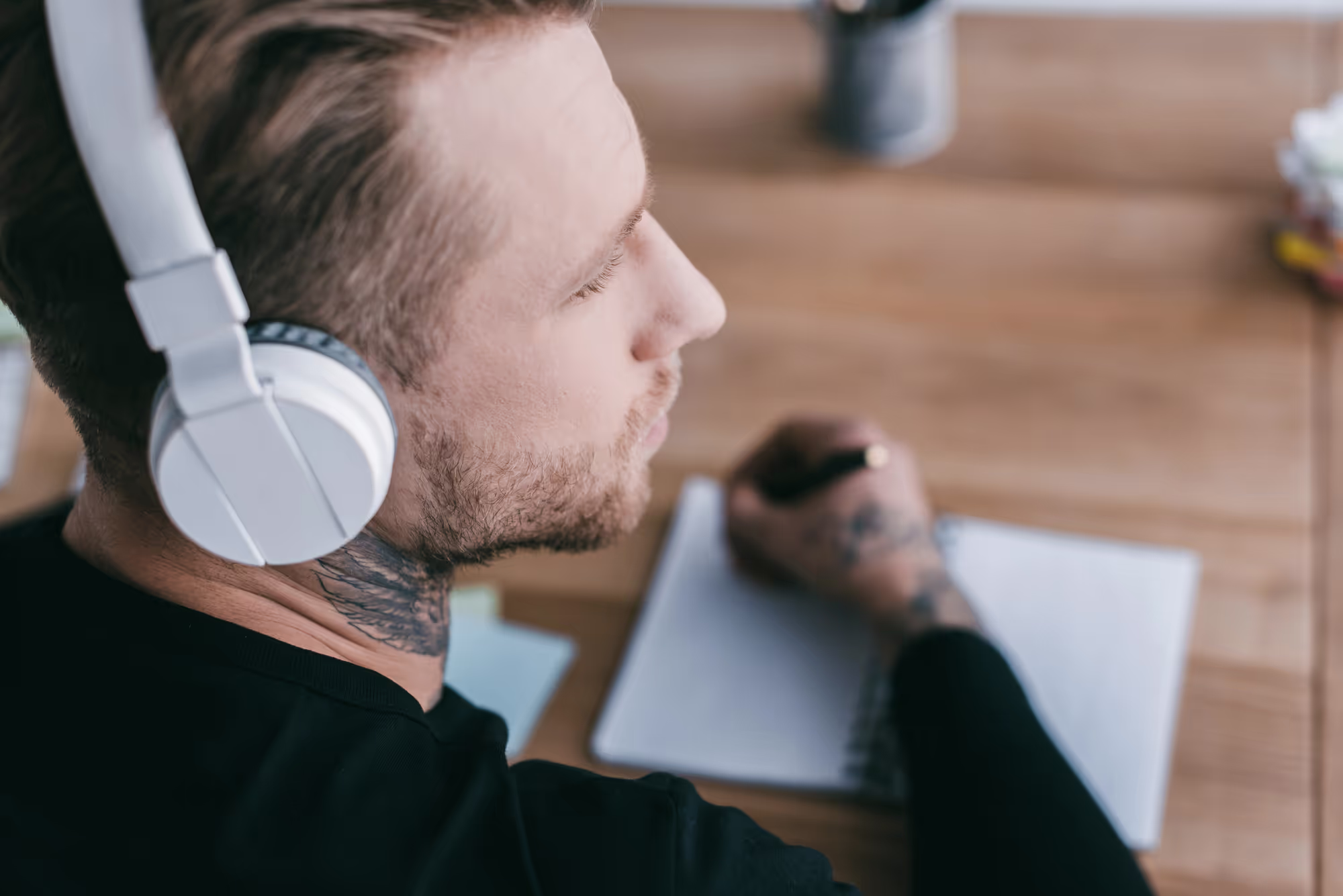 Close-up view of young businessman in headphones looking away while taking notes at workplace