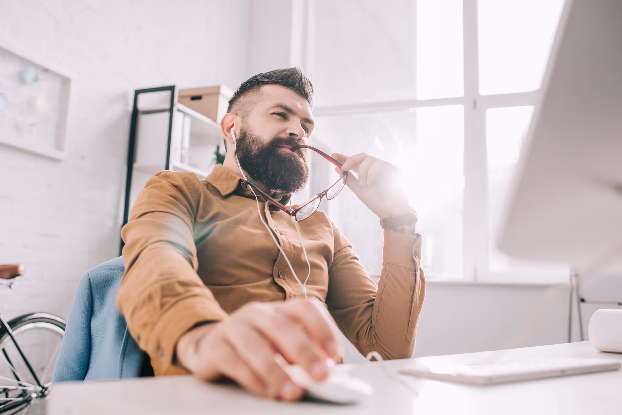 Pensive bearded adult businessman in earphones sitting and working at computer desk in office
