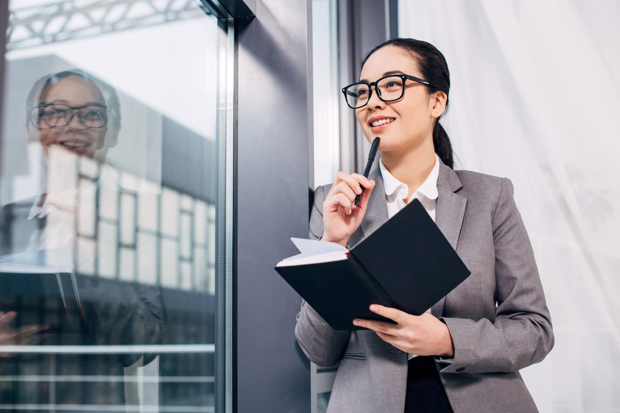 Pretty businesswoman smiling, standing by window and holding notebook and pen