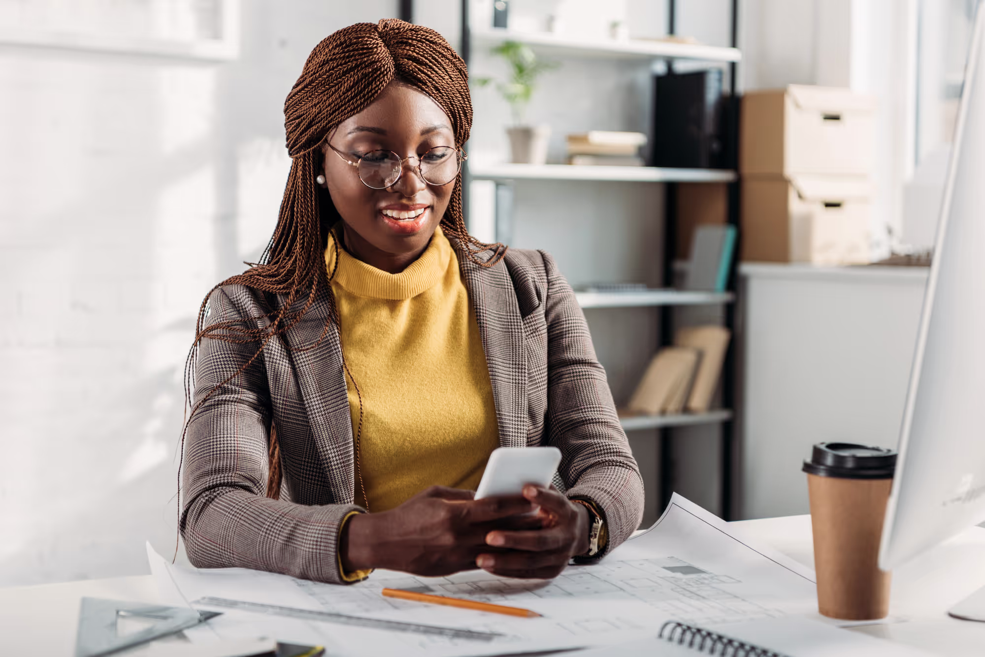Smiling african american female architect in glasses using smartphone and working at desk with blueprints in office