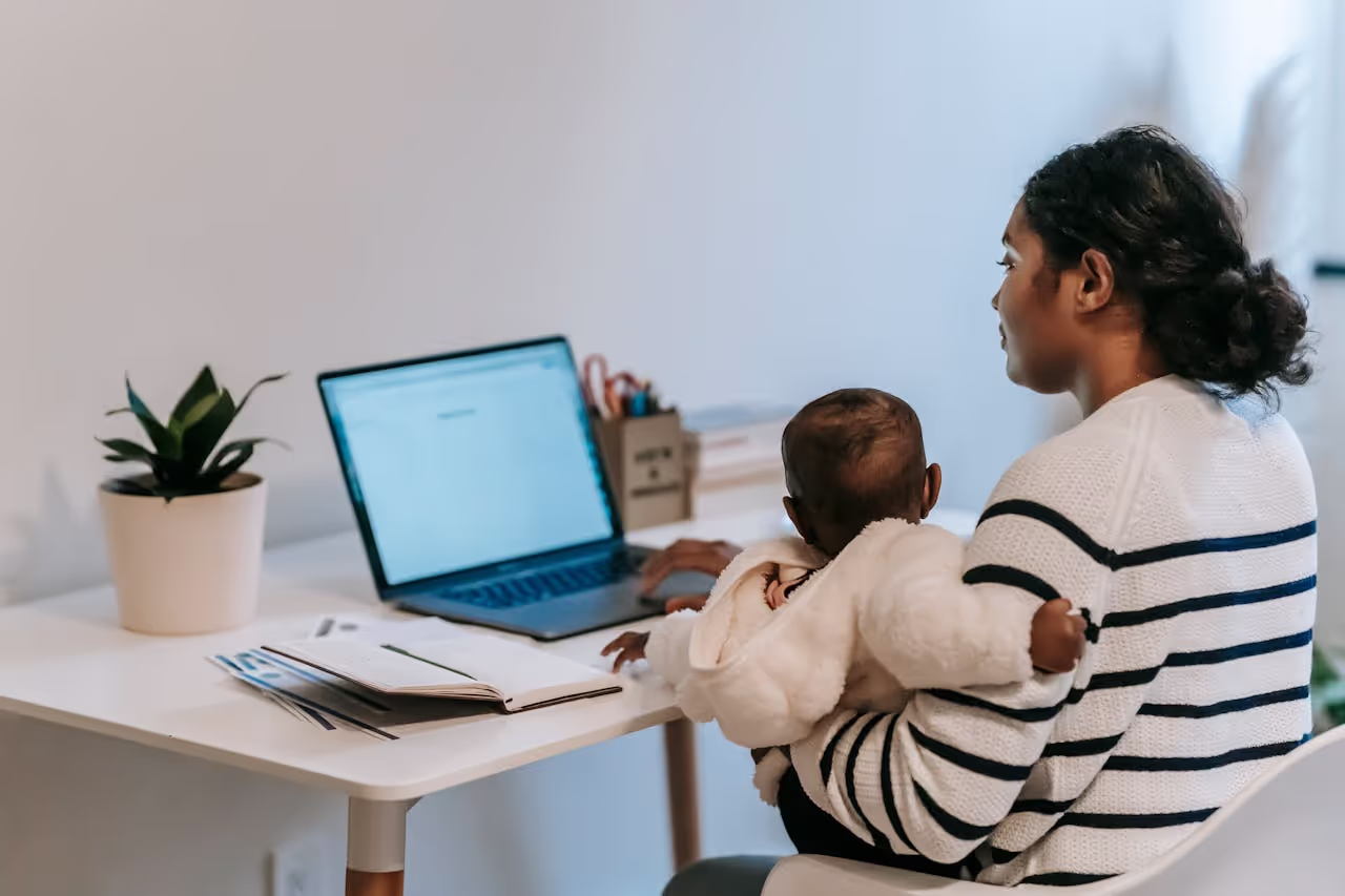 Support Student Parents College – Student Mother Balancing Studies and Childcare Student mother working on school assignments in front of a laptop while holding her baby, showing how colleges support student parents college success.