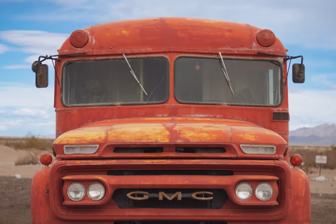A close-up, front view of a weathered, rusty orange GMC bus or truck in a desolate, arid landscape. The vehicle has two large windshields with wipers, round headlights, and a 'GMC' emblem on the grille. The paint is faded and peeling, showing patches of rust. In the background, there's a blue sky with scattered clouds and faint outlines of mountains.