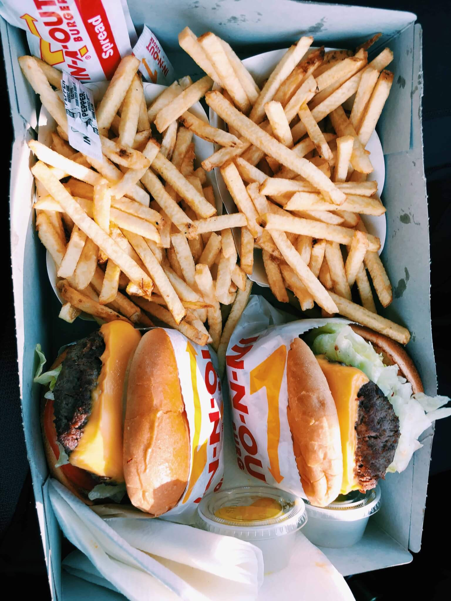 Overhead view of an In-N-Out Burger takeout box holding two cheeseburgers wrapped in branded paper, two generous servings of French fries, and small sauce cups with napkins.