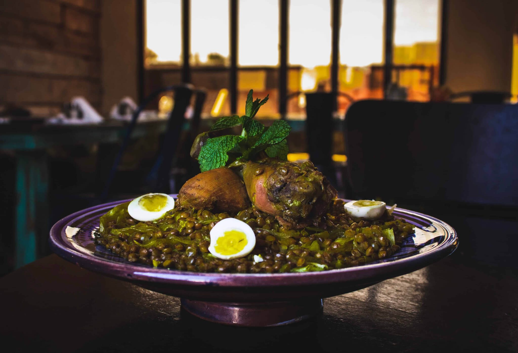 A plated dish of braised meat on a bed of lentils, garnished with mint and halved boiled eggs, served on a purple pedestal plate in a dimly lit restaurant.