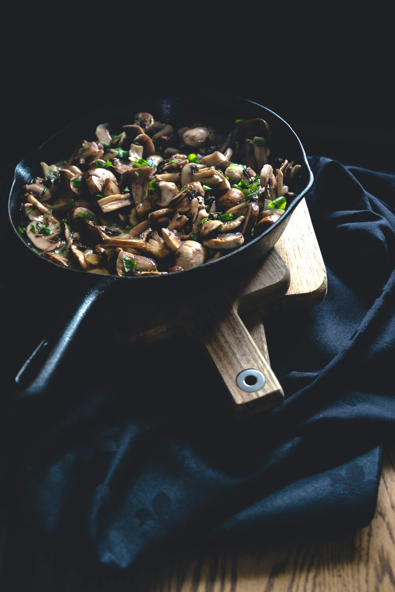 Cast-iron skillet filled with sautéed mushrooms and herbs on a wooden board against a dark background.