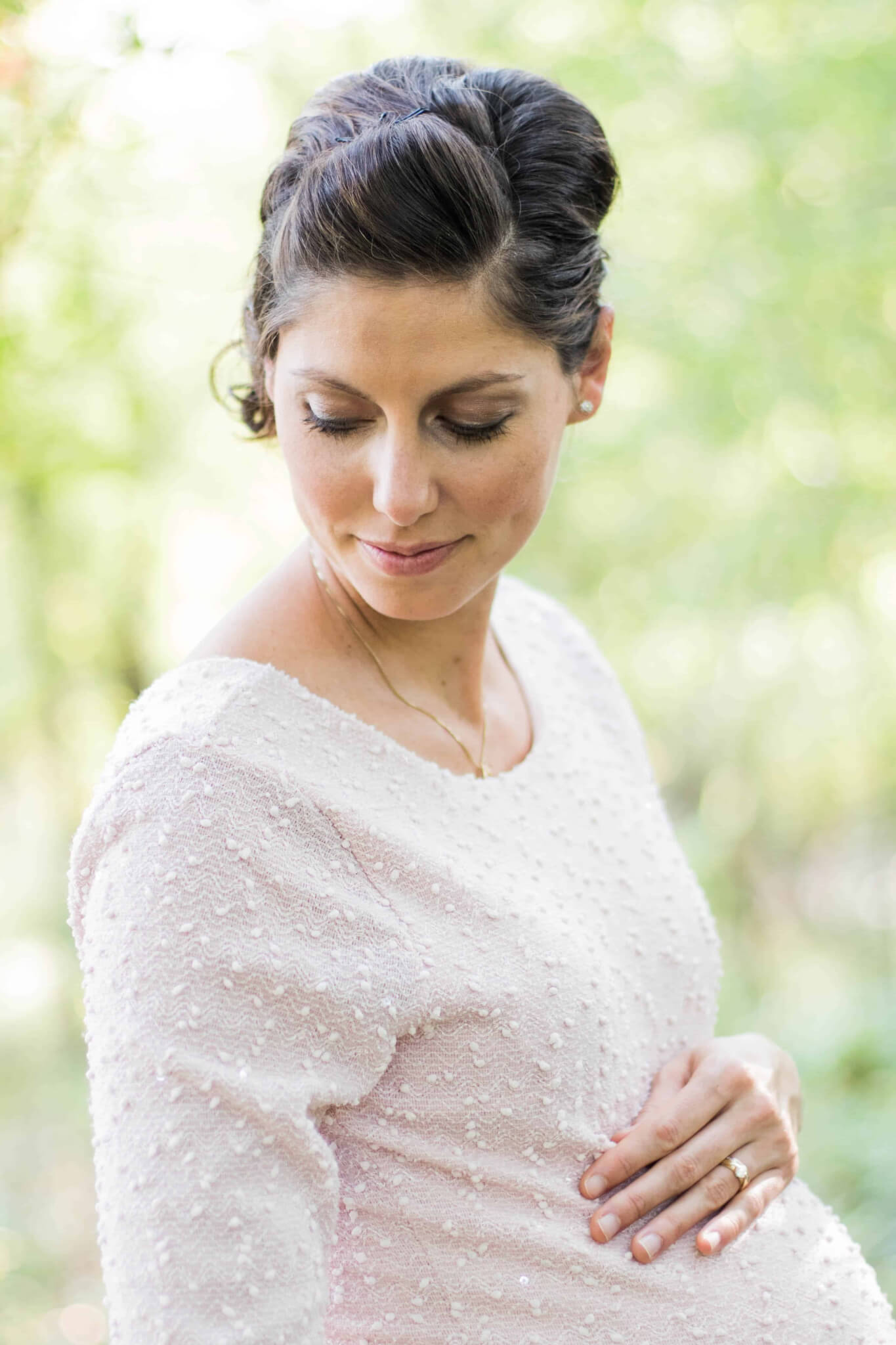 Close-up portrait of a pregnant woman in a light pink textured dress looking down with a gentle smile, resting a hand on their abdomen, set against a soft green, sunlit outdoor background.
