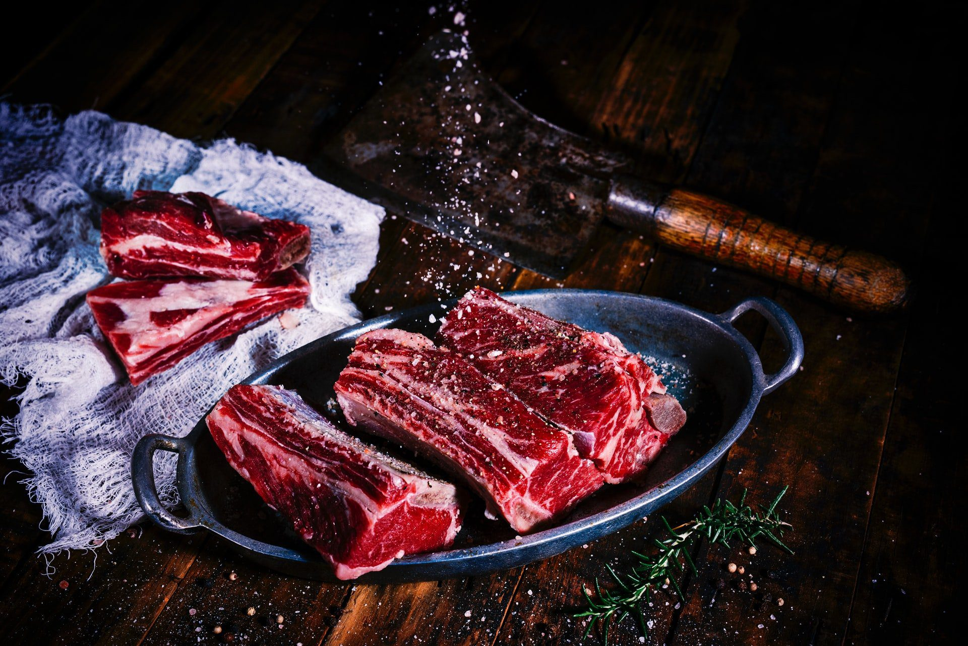 Raw beef short ribs in an oval metal pan on a rustic wooden table, being sprinkled with coarse salt, with a meat cleaver, cheesecloth, rosemary, and peppercorns nearby.