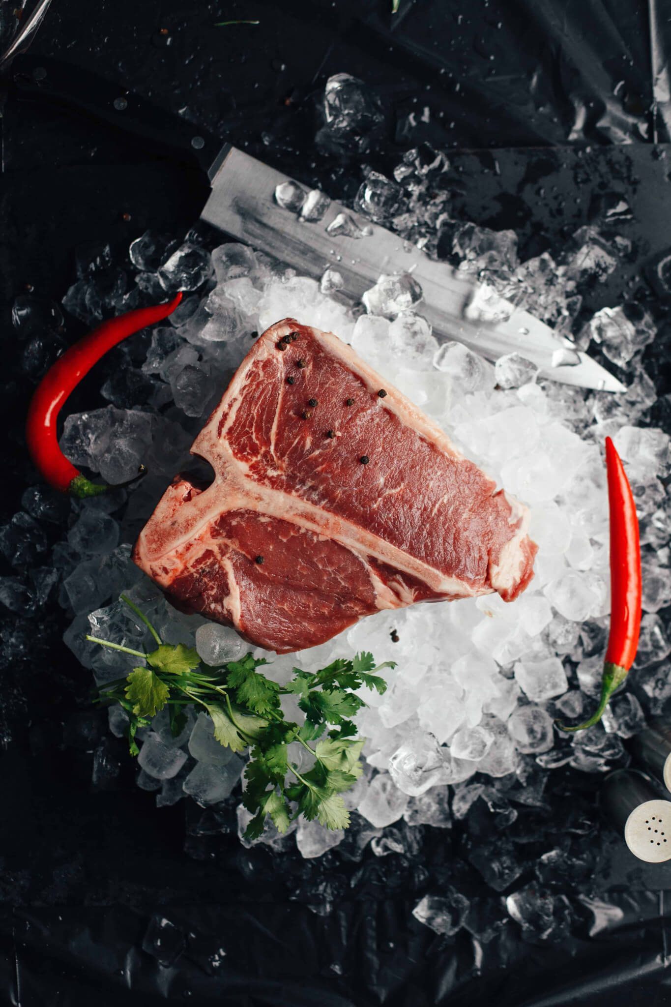 Raw T-bone steak on crushed ice with peppercorns, flanked by red chili peppers and cilantro, beside a large knife on a dark background.