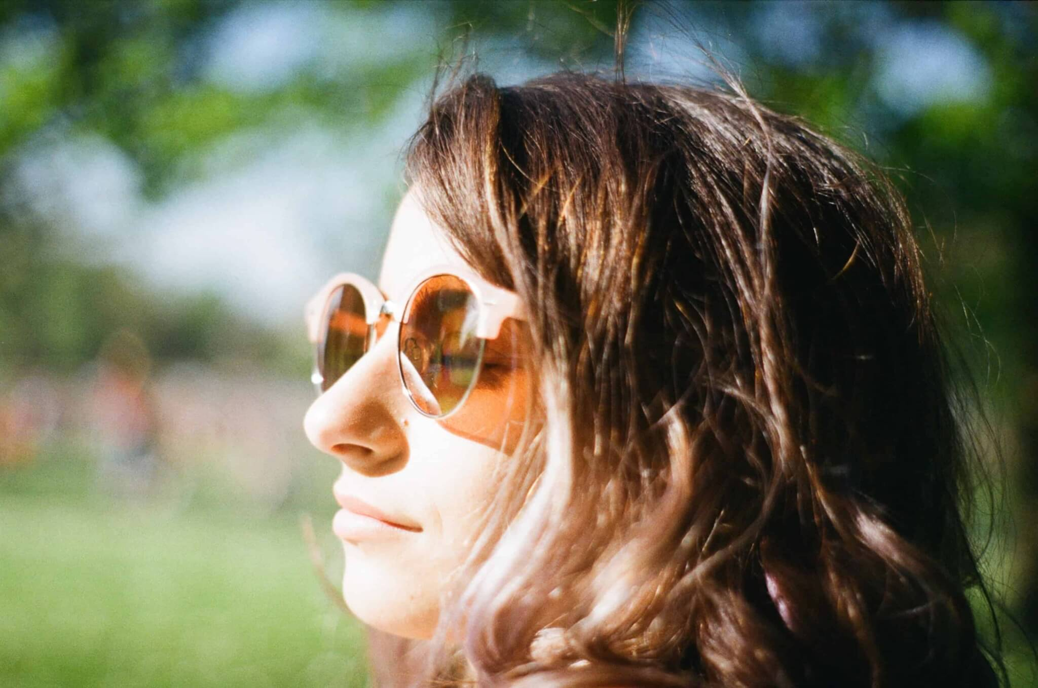 Close-up side profile of a person with long hair wearing round orange-tinted sunglasses outdoors, with soft green bokeh in sunlight.