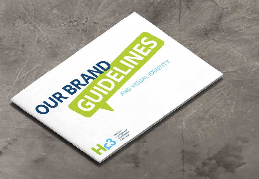 A white brand guidelines booklet titled 'OUR BRAND GUIDELINES AND VISUAL IDENTITY' with 'OUR BRAND' in blue, 'GUIDELINES' in white on a green speech bubble, and 'AND VISUAL IDENTITY' in light blue. The bottom left features the Hc3 logo in green and blue, with 'Healthy Campus and Community Coalition' below it. The booklet rests diagonally on a dark gray, textured surface.