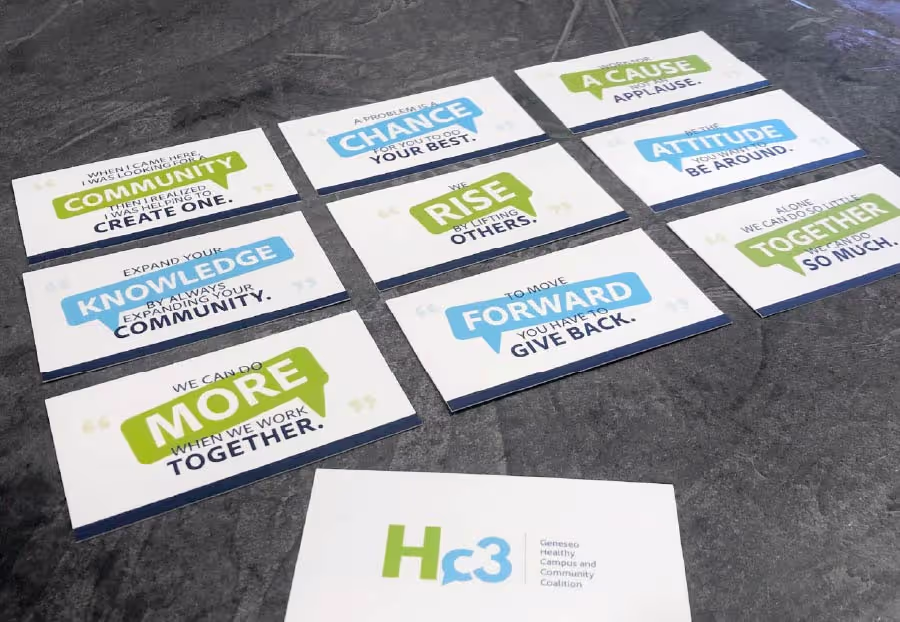 Ten white cards are spread across a dark grey surface. Nine cards feature motivational quotes with key words highlighted in green or blue speech bubble shapes, such as 'WHEN I CAME HERE, I WAS LOOKING FOR A COMMUNITY THEN I REALIZED I WAS GOING TO CREATE ONE' and 'WE CAN DO MORE WHEN WE WORK TOGETHER'. The tenth card, in the foreground, displays the logo 'Hc3' in green and blue, with 'Geneseo Healthy Campus and Community Coalition' written below it.