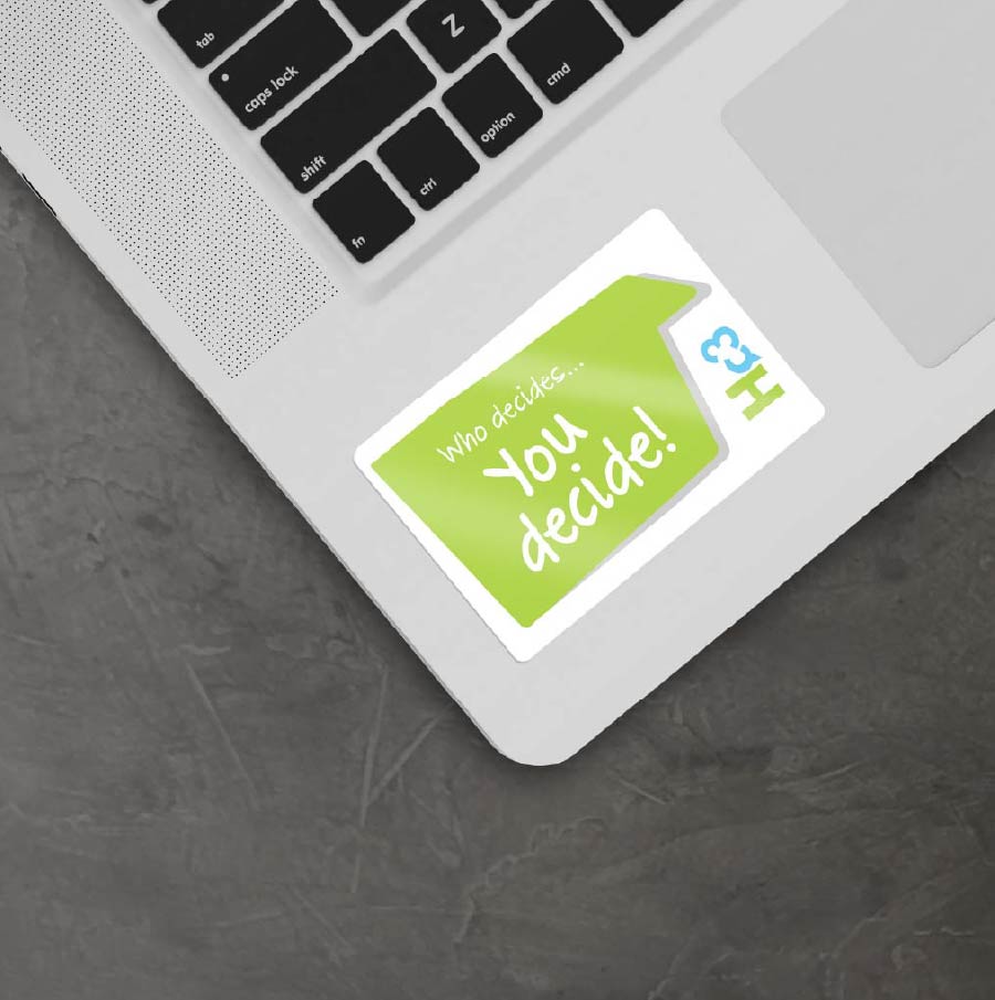 A green speech bubble sticker with white text that reads 'Who decides... You decide!' and a small H3 logo in blue and green, placed on the white palm rest of a laptop. The keyboard is partially visible at the top, and a dark, textured surface is in the background.