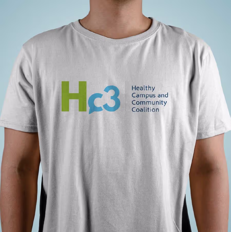 A person wearing a white t-shirt with a logo and text. The logo on the left features a large green 'H' followed by a light blue 'c' shaped like a speech bubble, and then a light blue '3'. To the right of the logo, the words 'Healthy Campus and Community Coalition' are printed in dark blue text, stacked on three lines. The person's neck and shoulders are visible against a light blue background.