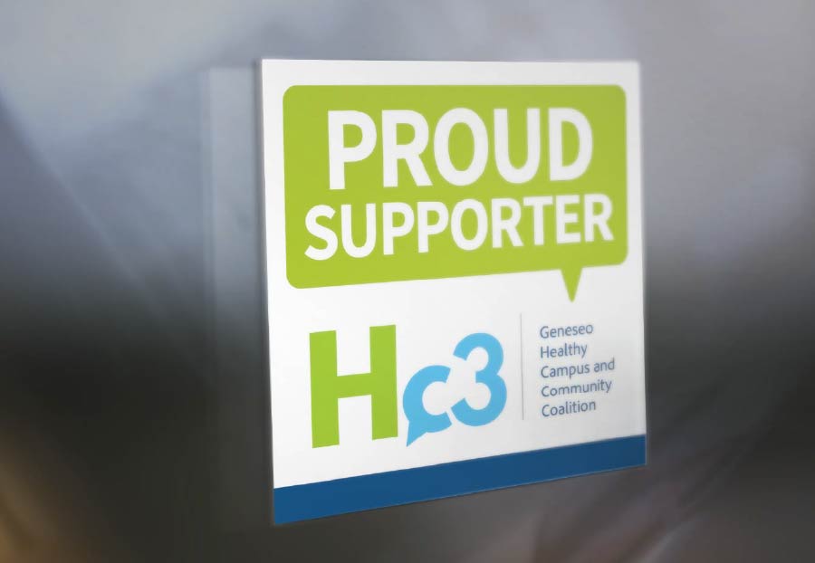 A white sign with a green speech bubble at the top that reads 'PROUD SUPPORTER'. Below that, in larger font, is 'Hc3' with the 'c' formed by a blue speech bubble containing the number 3. To the right of 'Hc3' is smaller text that reads 'Geneseo Healthy Campus and Community Coalition'. A blue strip runs along the bottom of the sign. The sign is angled against a muted, out-of-focus background, casting a shadow.