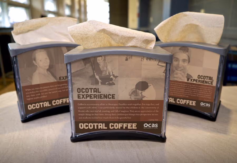 Three tissue dispensers on a table, each featuring images and text promoting 'Ocotal Coffee' and 'Ocotal Experience'. The central dispenser, most in focus, shows a woman and child with a story about community coffee in Nicaragua.