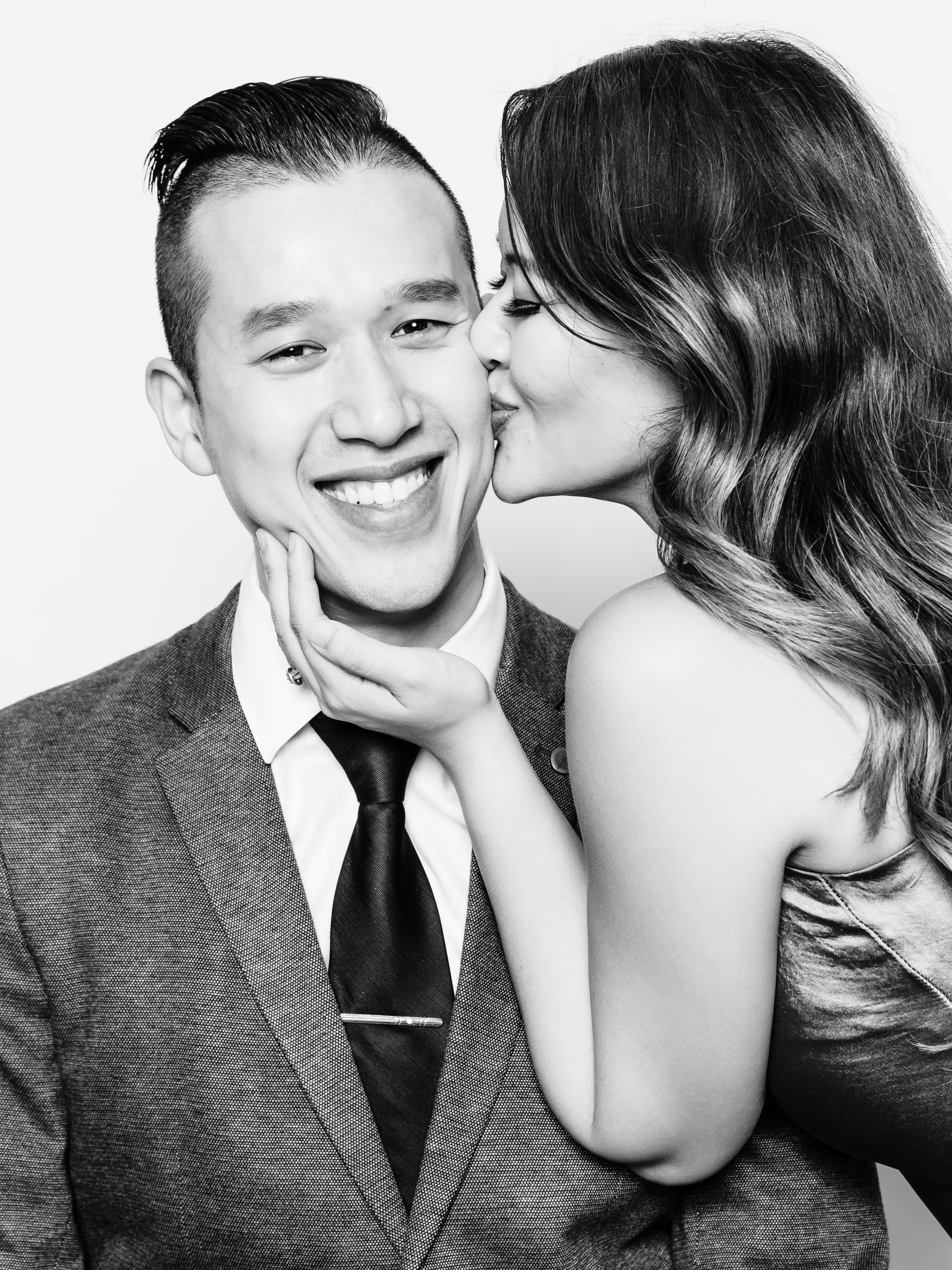 Wife holding on to her husband's chin while she kisses his cheek from the right side while the husband is facing the camera grinning in a black & white photo