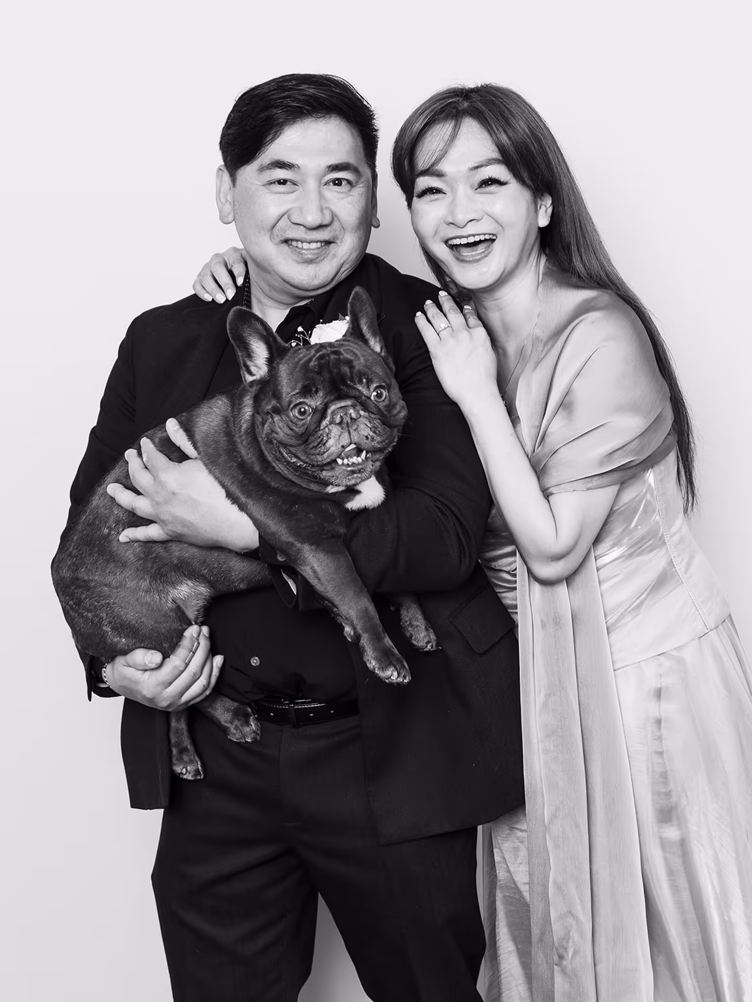 An Asian couple facing the camera while the wife holds on to the husband on the right side while the husband is holding on to their black dog in a black & white photo
