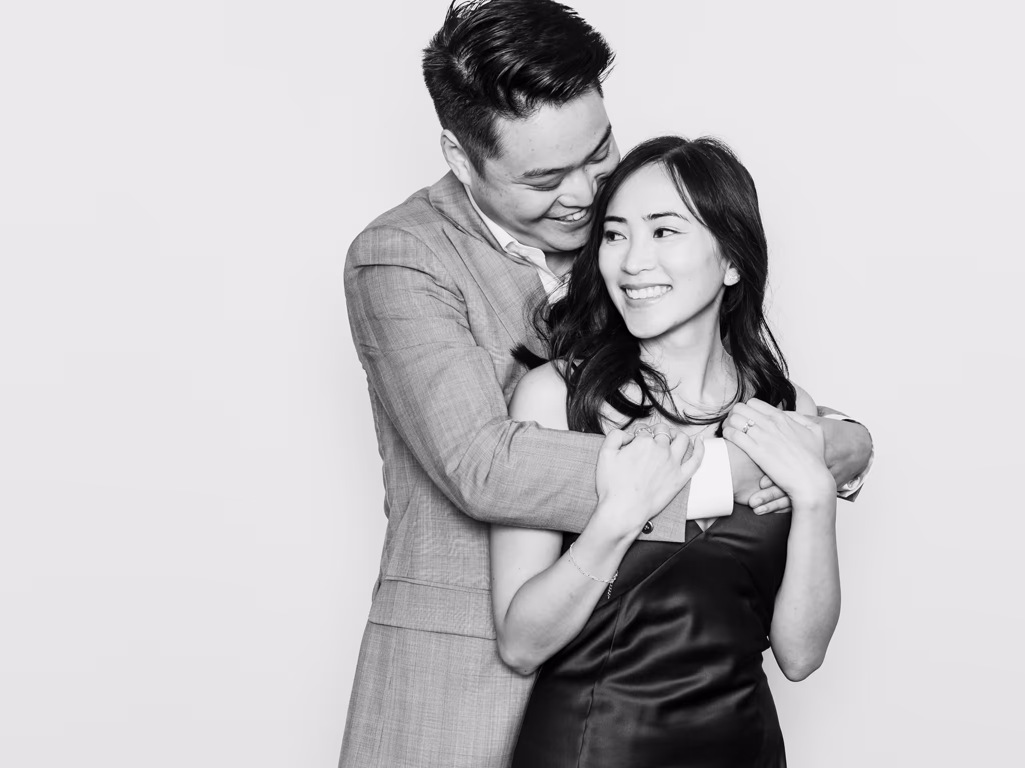 Asian man hugging her wife from behind in a black and white photo while she holds her husband's hand